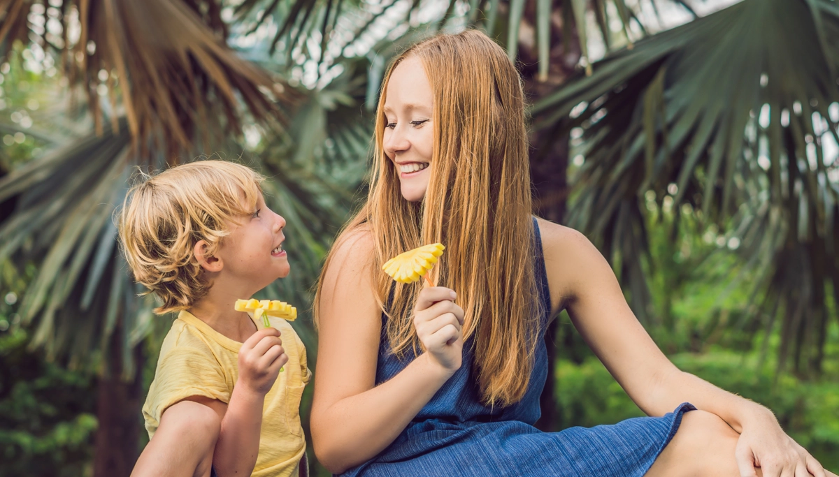 A smiling mother and son sitting outdoors in front of tropical palm trees, while holding pineapple slices on sticks